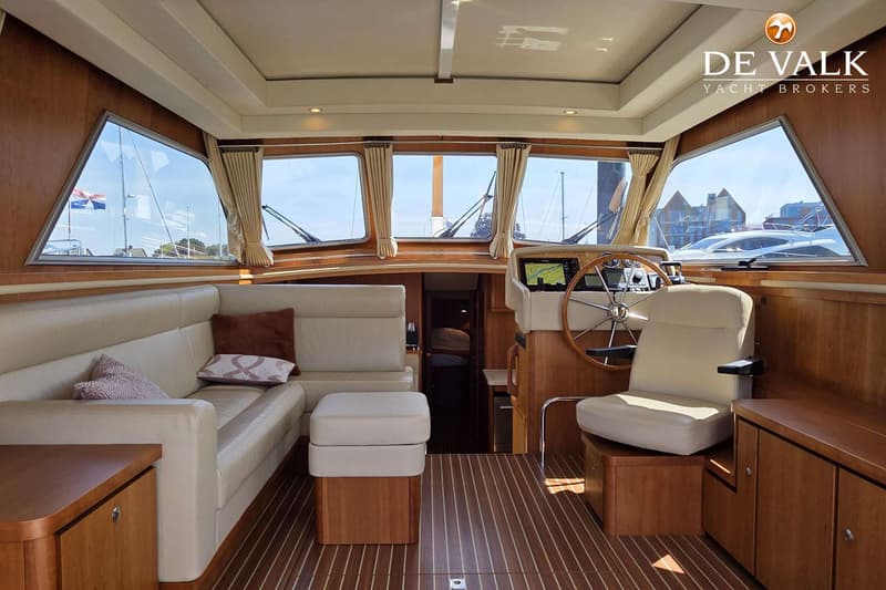 De foto toont het interieur van een boot met een stuurstand, een beige bank, houten vloer en ramen met uitzicht naar buiten.