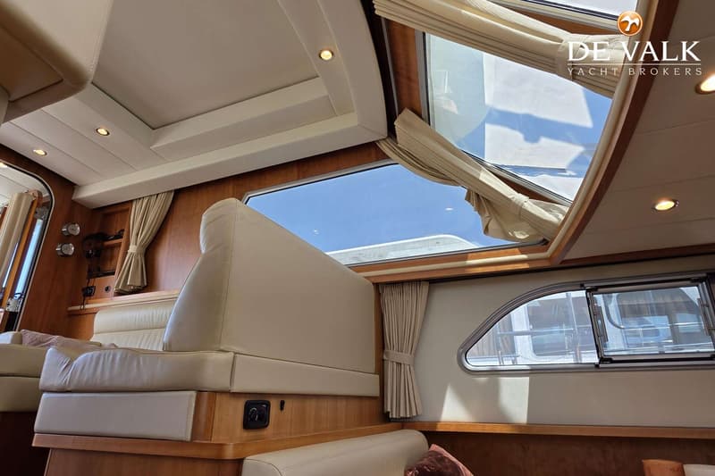 De foto toont het interieur van een boot met beige bekleding, houten panelen, ramen met gordijnen en een open dakluik.