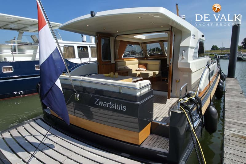 Een motorjacht met de naam "Zwartsluis" ligt afgemeerd aan een houten steiger, met een Nederlandse vlag aan de achtersteven.