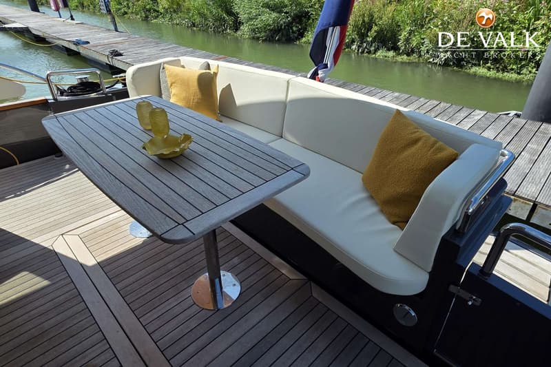 Een houten tafel met twee glazen en een schaal staat voor een witte bank met gele kussens op een bootdek.