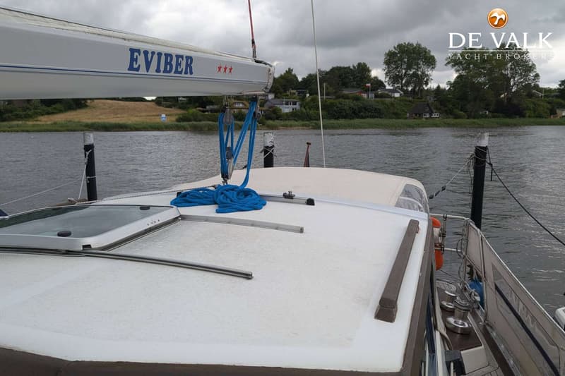 Een witte boot met de naam "EVIBER" en drie rode sterren op de giek, ligt aangemeerd aan een waterweg. — LM 35 Vitesse