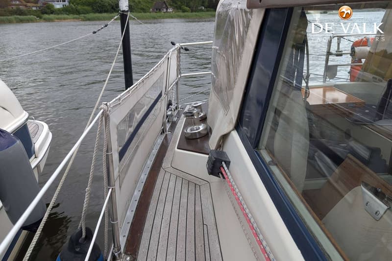 Een zijaanzicht van een boot met een houten dek, reling, touwen en een afdekzeil, liggend aan het water. — LM 35 Vitesse