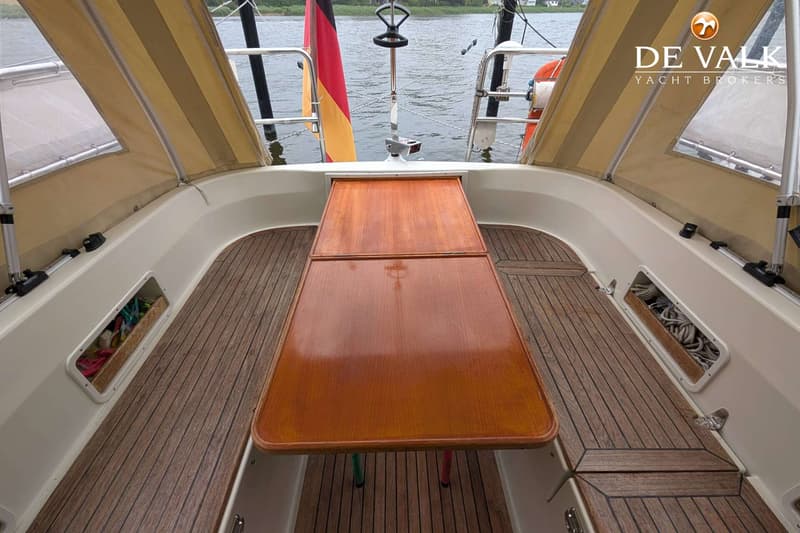 Een houten tafel met twee bladen staat op een teakhouten dek, omringd door een beige kuiptent en de Duitse vlag.