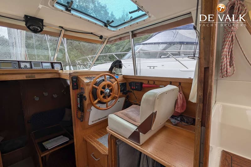 De foto toont de stuurstand van een boot met een houten stuurwiel, een beige stoel en diverse navigatie-instrumenten.