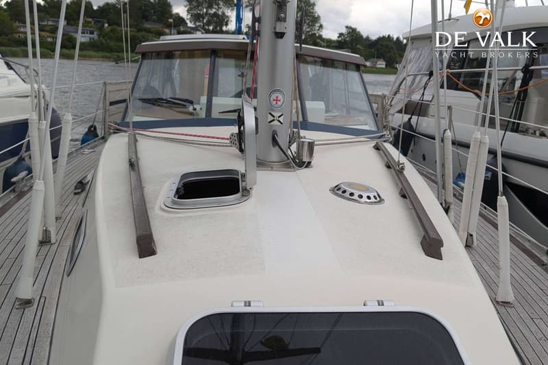 Een dek van een boot met een mast, luiken, en een kajuit met ramen, met op de achtergrond water en begroeiing.
