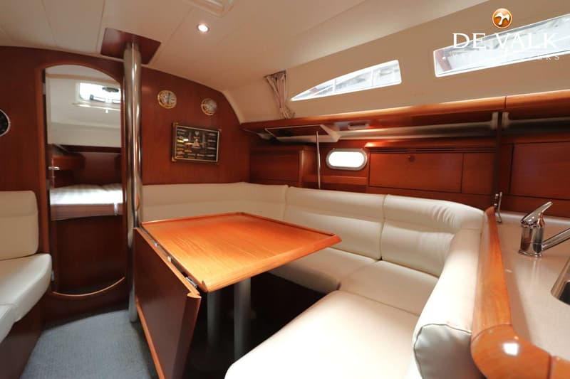 De foto toont een interieur van een boot met een houten tafel, witte banken, houten kasten en een doorgang naar een…