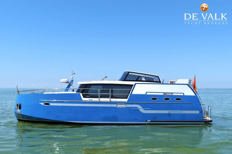 Een blauw-witte motorboot met de naam "THOMASZ 41" is te zien op het water met windmolens op de achtergrond.
