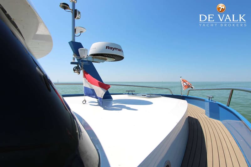 Een deel van een boot met een Raymarine radar, twee vlaggen, een reling en een houten dek is zichtbaar.