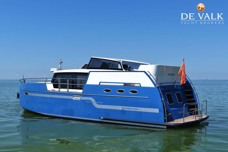 Een blauw-witte motorboot met de naam "THOMASZ 2.41" ligt op het water, met windmolens op de achtergrond.