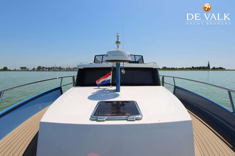 Een witte boot met een blauwe romp en een Nederlands vlaggetje vaart op een meer met een jachthaven en een dorp op de…