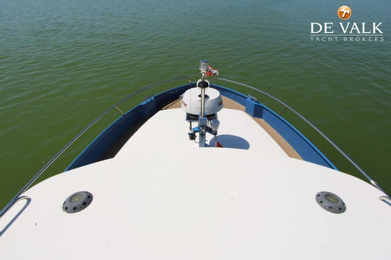 De boeg van een witte boot met blauwe rand en houten dek, met een radar en navigatielichten, vaart op groen water.