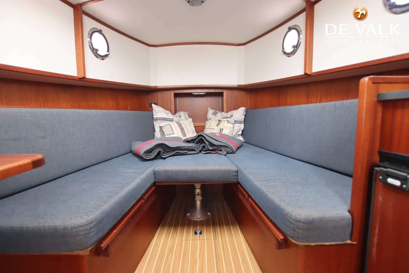 Een interieur van een boot met een U-vormige bank met blauwe kussens, twee patrijspoorten en een houten vloer.