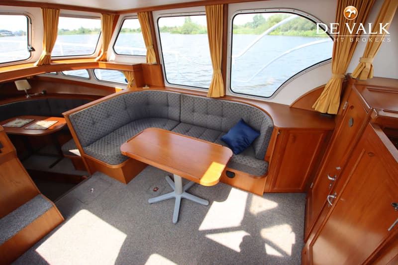 De foto toont het interieur van een boot met een zithoek, houten meubels, ramen met gele gordijnen en uitzicht op water.