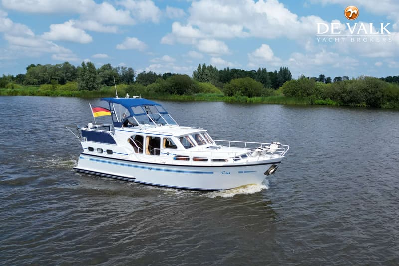 Een witte motorboot met blauwe accenten vaart op een waterweg met groene oevers onder een bewolkte hemel. — Pedro Skiron 35