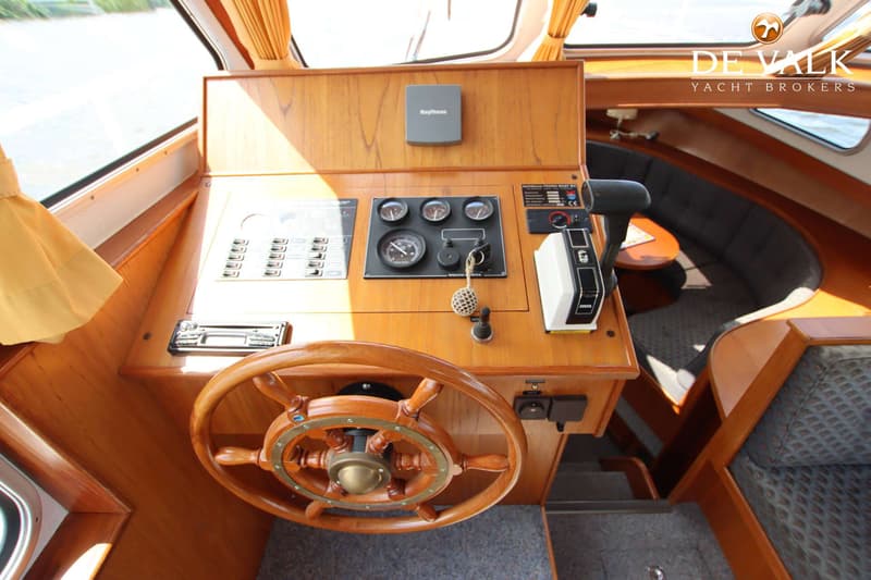 Een houten stuurwiel en dashboard met meters, schakelaars en een Raytheon-apparaat zijn zichtbaar in de stuurhut.