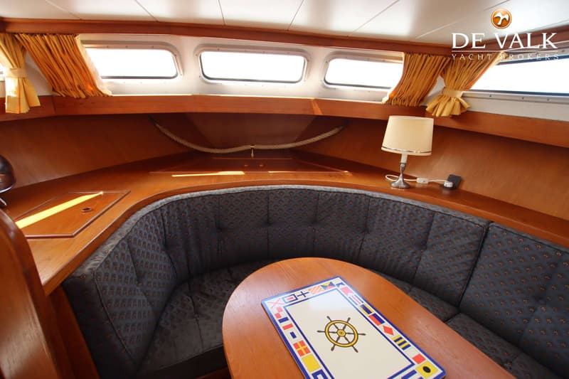 Een kajuit met een ronde zithoek, een houten tafel met een nautisch motief, ramen met gele gordijnen en een lamp.