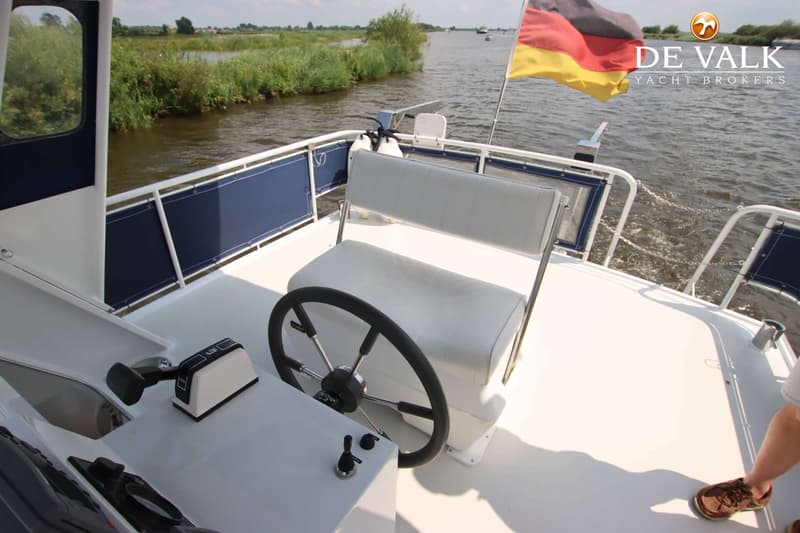 Een stuurstand met een wit bankje en een zwart stuur op een boot, met een Duitse vlag en water op de achtergrond.
