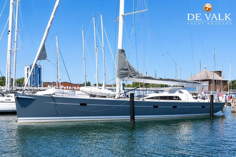 Een grijze zeilboot met witte opbouw ligt afgemeerd in een haven, met andere boten en gebouwen op de achtergrond.