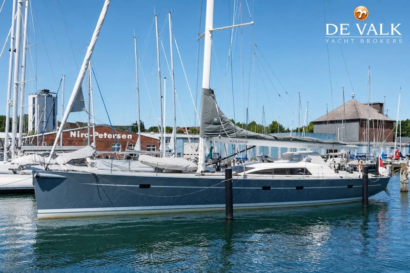 Een grijze zeilboot met witte mast en grijze zeilhoes ligt in het water, met gebouwen en andere boten op de achtergrond.