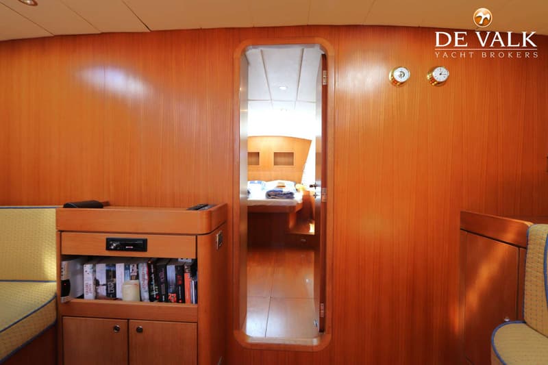 Een houten interieur van een boot met een open deur naar een slaapcabine, een boekenkast en twee klokken aan de muur.