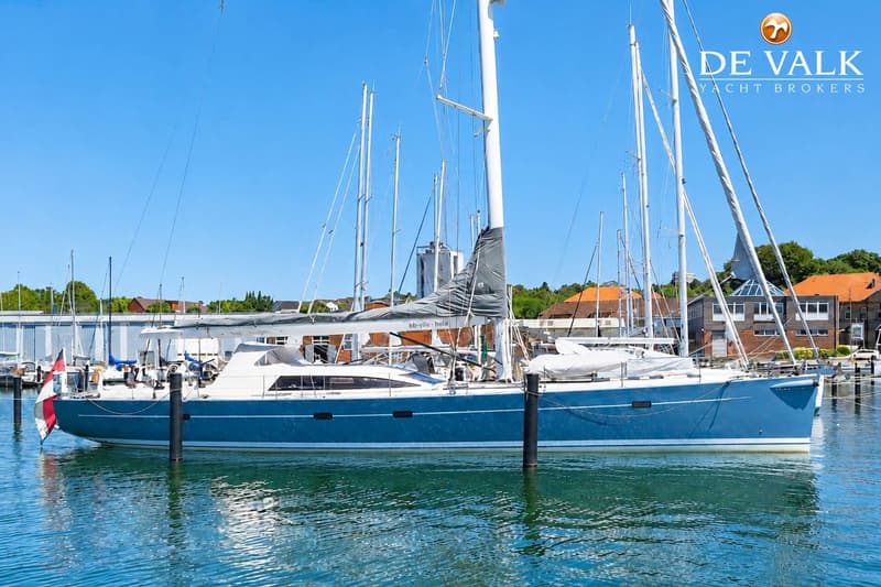 Een blauwe zeilboot met witte opbouw ligt afgemeerd in een haven, met andere boten en gebouwen op de achtergrond.