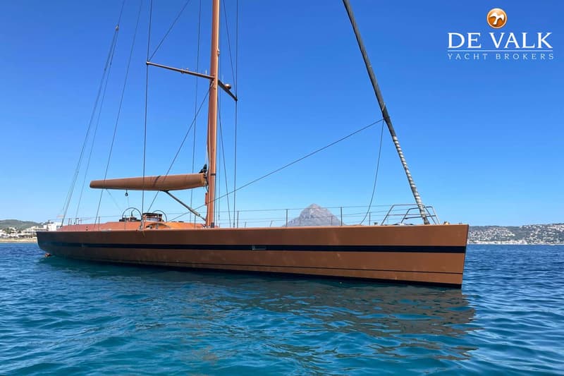 Een bruine zeilboot met twee masten en een zwarte streep op de romp drijft op blauw water onder een heldere lucht. — Wally 88