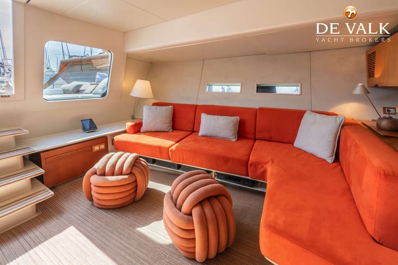 Een interieur van een boot met een oranje L-vormige bank, twee oranje poefs, een tablet en een raam met uitzicht op een…