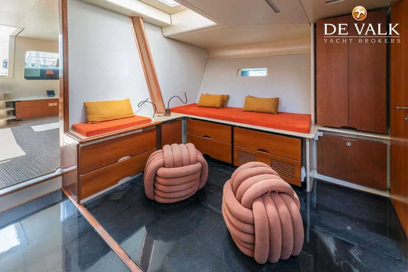 De foto toont een kajuitinterieur met oranje kussens op banken, houten kasten en twee roze poefs op een donkere vloer.