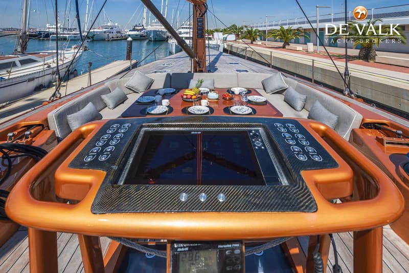 Een dek van een zeiljacht met een tafel, zitbanken, een navigatiescherm en een fruitmand, liggend in een haven.