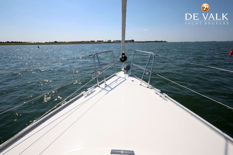 De boeg van een witte zeilboot met een opgerold zeil en reling, varend op water met land in de verte. — Bavaria 37 Cruiser