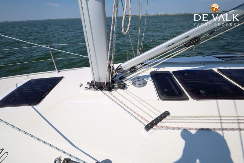 Een wit dek van een zeilboot met mast, zeil, lijnen, luiken en een Selden giek, varend op water met windmolens op de…