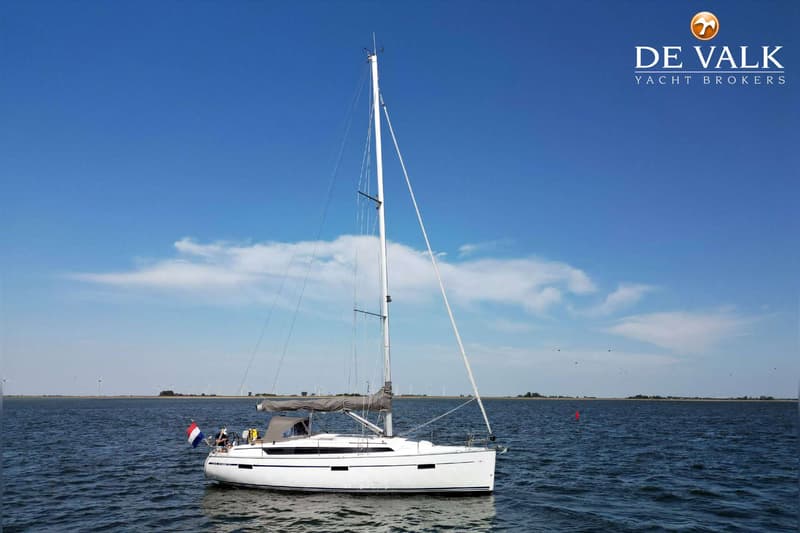 Een witte zeilboot met een grijze huik en een Nederlandse vlag vaart op kalm water onder een blauwe lucht.