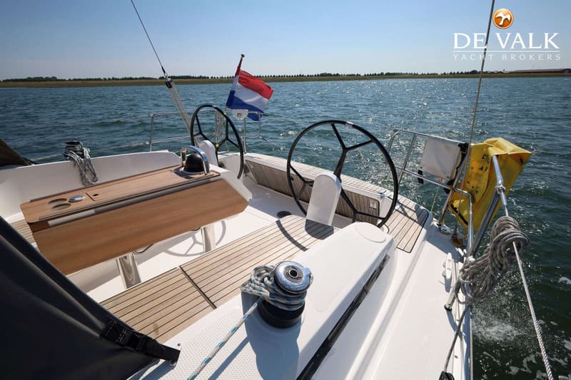 Een achterdek van een zeilboot met twee stuurwielen, een houten tafel en een Nederlandse vlag, varend op water.