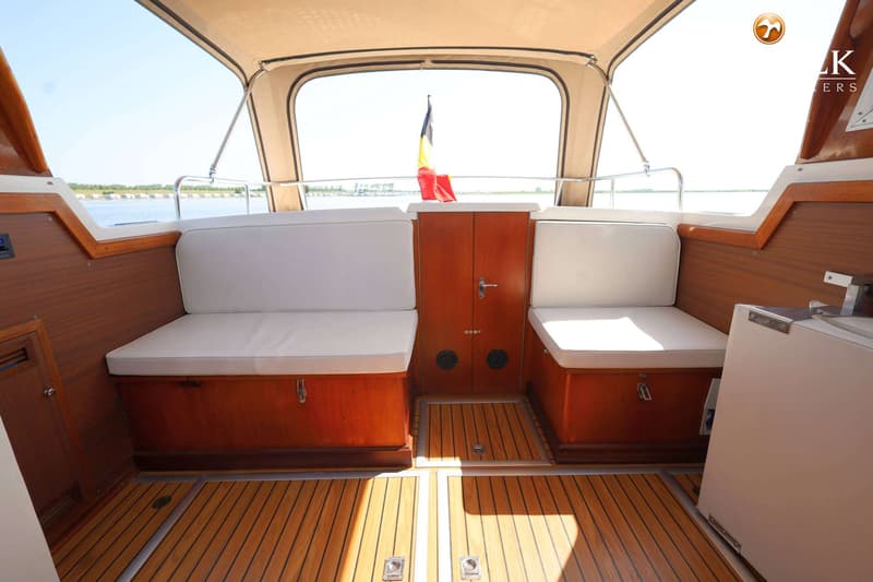 Een interieur van een boot met houten vloer en wanden, witte kussens en een Belgische vlag aan de achterzijde.