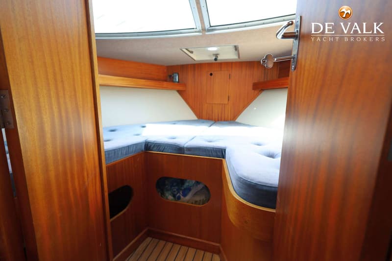 De foto toont het interieur van een boot met een V-vormig bed met blauwe kussens, houten wanden en een luik in het plafond.