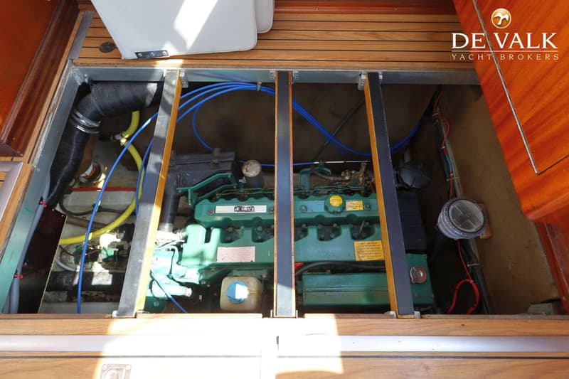 Een groene scheepsmotor met blauwe slangen en gele leidingen is zichtbaar onder een houten vloer.