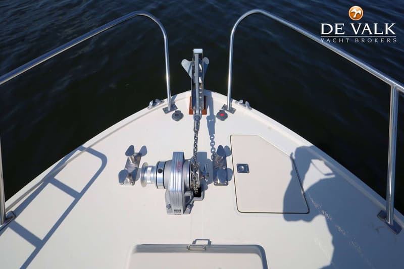 De boeg van een boot met een ankerlier, anker, ketting en reling, met donker water op de achtergrond.
