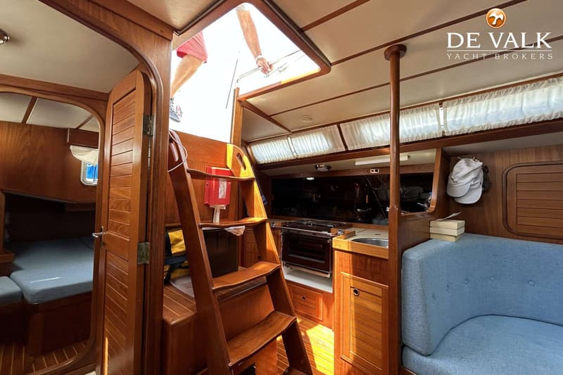 Een interieur van een boot met houten afwerking, een trap, een kombuis, een blauwe bank en een persoon die uit een luik…