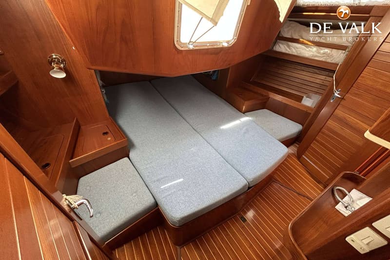Een kajuit met houten wanden en vloer, een bed met grijze kussens en een raam met een luik. — Maestro 35
