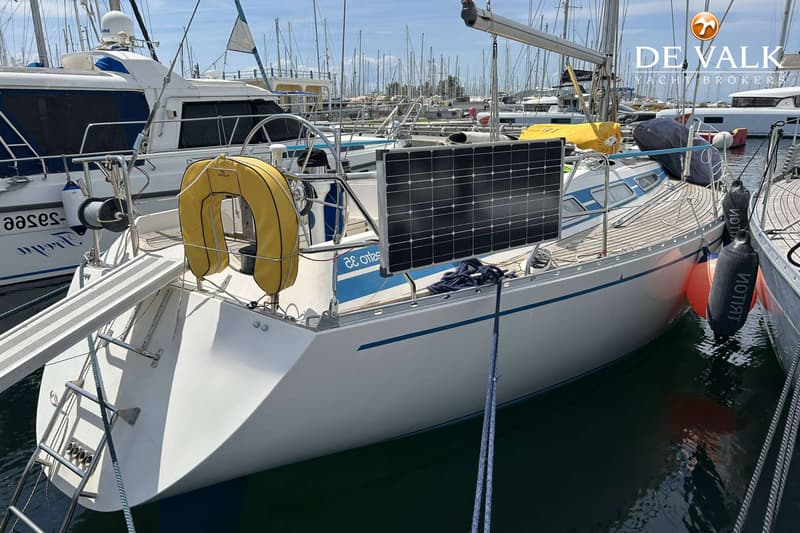 Een witte zeilboot met blauwe strepen en een zonnepaneel ligt afgemeerd in een haven vol met andere boten. — Maestro 35