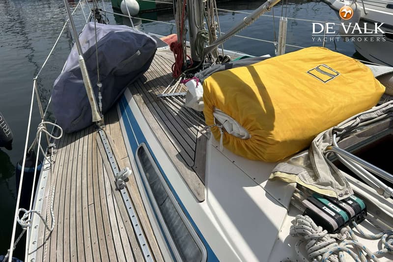 Een bovenaanzicht van een bootdek met houten planken, een blauwe streep, en twee zeilhoezen in grijs en geel.