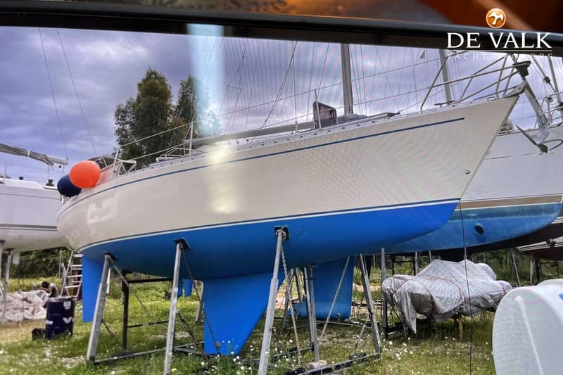 Een zeilboot met een crème romp en blauwe onderkant staat op bokken in het gras, met bomen op de achtergrond. — Maestro 35