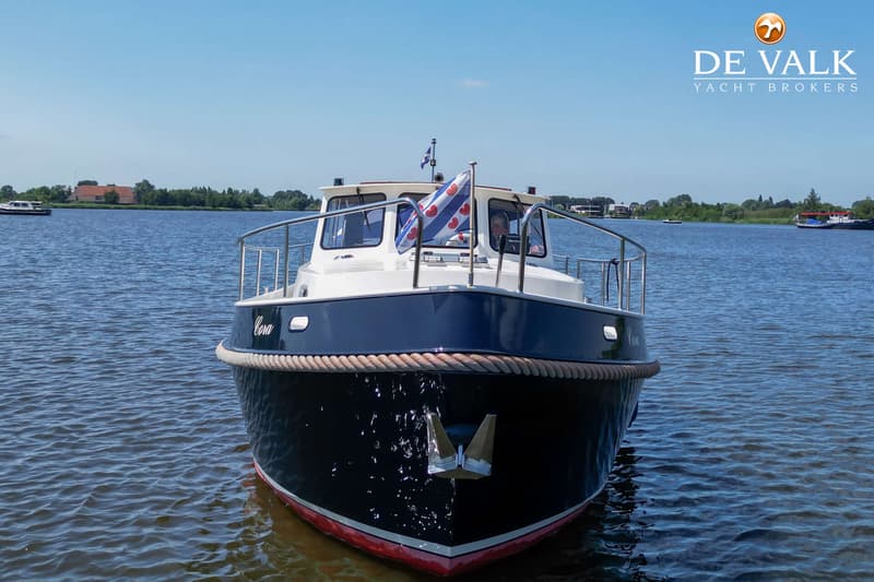 Een donkerblauwe boot met witte opbouw en een rood-witte onderkant ligt in het water, met een Friese vlag op de boeg.