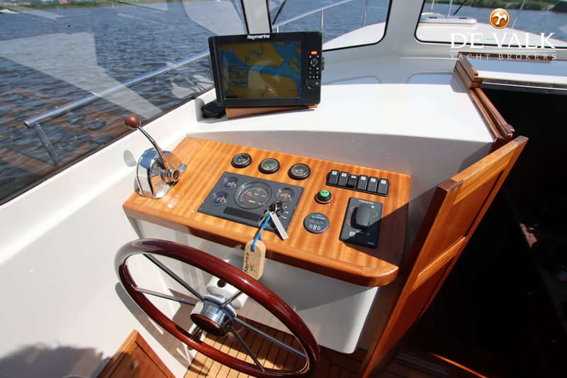 Een stuurstand met een houten stuurwiel, diverse meters, schakelaars, een Raymarine navigatiescherm en een gashendel.