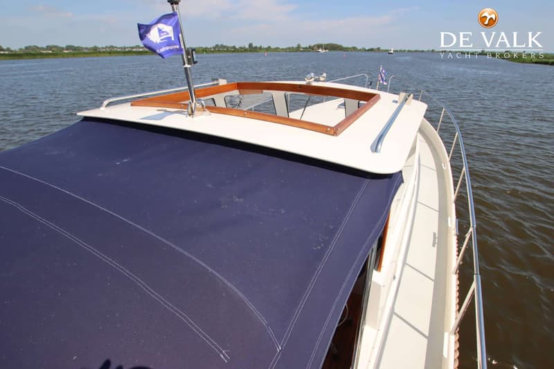Een boot met een blauw dekzeil en een vlag op de achtersteven, varend op water met land in de verte.