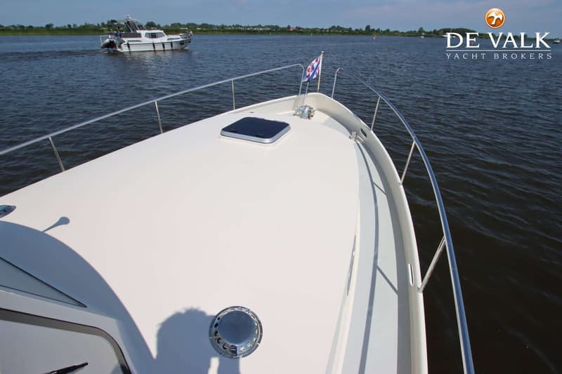 De boeg van een witte boot met een luik en een vlag, varend op water met een andere boot op de achtergrond.