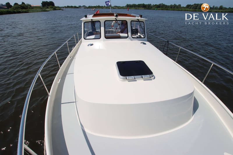 Een witte boot vaart op een waterweg met een persoon aan het roer en een open luik op het voordek.