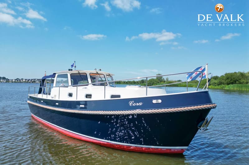 Een donkerblauwe boot met witte opbouw en rode onderkant, genaamd "Cora", ligt in het water onder een blauwe lucht.