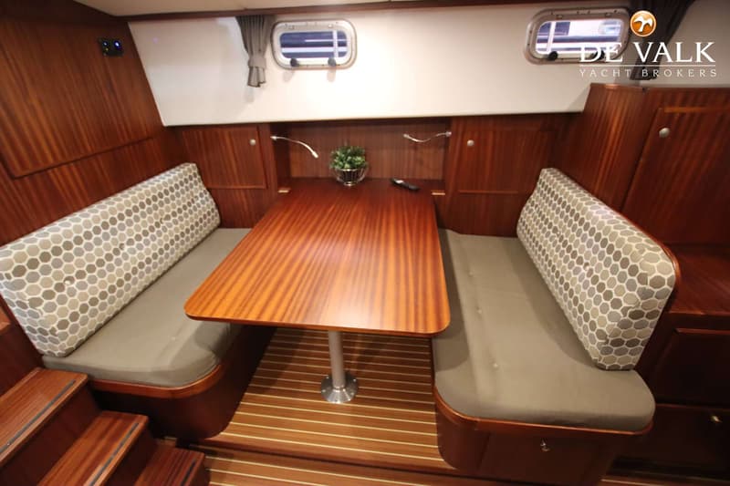 Een kajuit met een houten tafel, twee banken met kussens, houten wanden en vloer, en twee patrijspoorten.