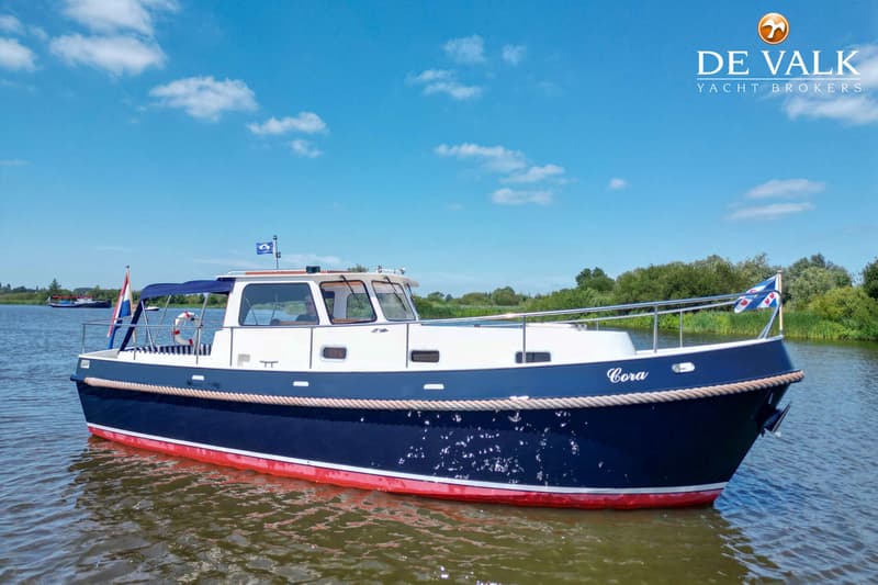 Een blauw-witte motorboot met rode onderkant en de naam "Cora" op de boeg vaart op een waterweg.